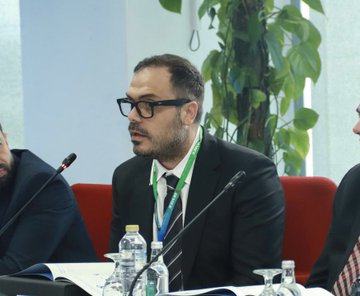 A bespectacled man in a dark suit and tie sits at what appears to be a panel or conference table, with a water bottle and microphone in front of him.