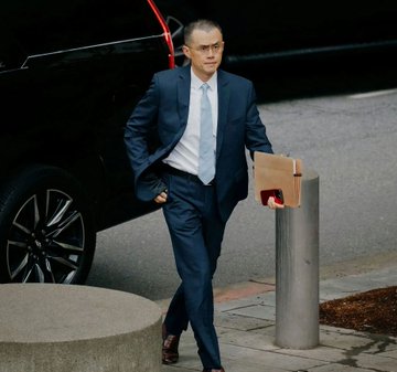 The image features a man dressed in a suit walking down a sidewalk while carrying a briefcase. The background includes a car parked nearby and another person standing further back along the street. The overall scene suggests that the man might be heading to or from work, possibly as an executive or businessman.