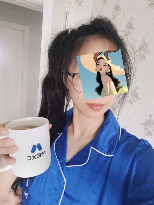 A selfie of a young woman wearing glasses and blue pajamas, holding a white mug with a 'MEXC' logo, with her face partially covered by an anime-style girl sticker.