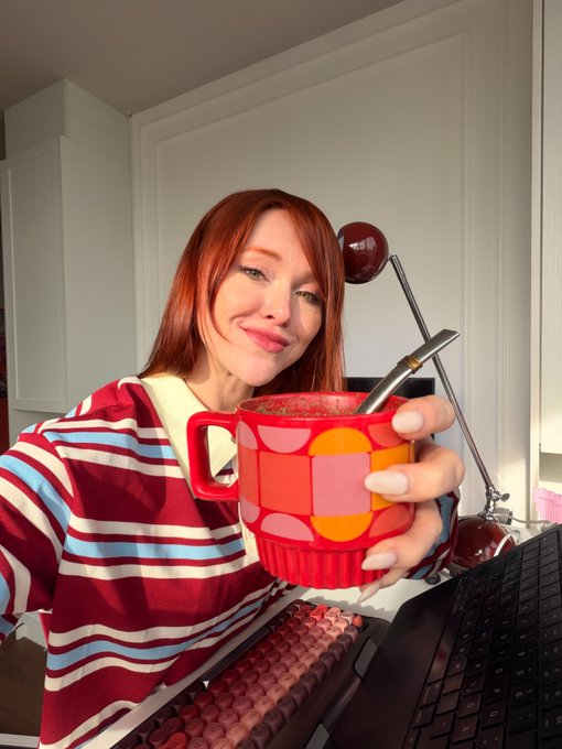 A selfie of a smiling woman with red hair holding a colorful patterned mug with a brush in it, positioned in front of a computer keyboard and a red desk lamp.