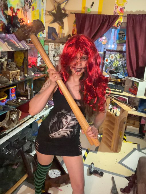 A red-haired artist poses in a colorful studio holding two wooden sledgehammers, matching the post’s “2 sledgehammers are better than 1” line for the 7 Days 10 Tez #walls event by @tornado_ilus. Her black tank top features the artwork from her self-portrait.