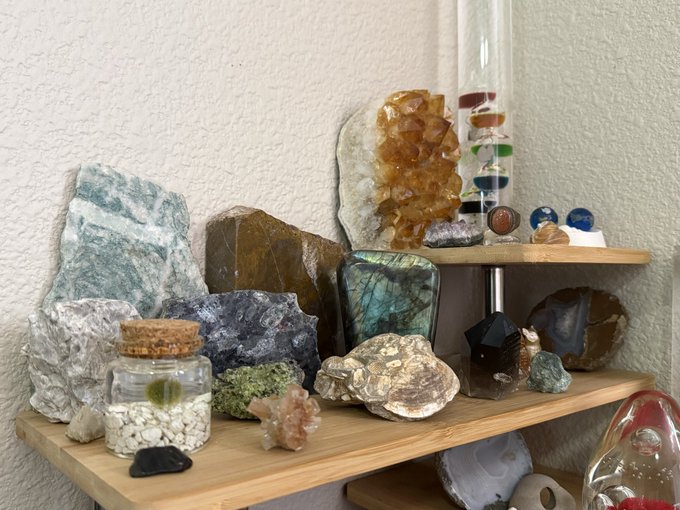 A collection of various rocks, minerals, and crystals displayed on wooden shelves, including a large citrine cluster, labradorite, and a jar with small stones.