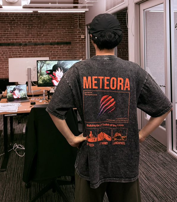 A person wearing a black cap stands with their back to the camera, showcasing a faded black t-shirt with bold orange "METEORA" lettering and graphics featuring the tagline "Building the LP Toolkit of the Future" alongside "DLMM," "ARMY," and "WINgMphos." In the background, an office setting with exposed brick walls and a computer monitor displaying a red bird character complements the accompanying post about Meteora as "the greatest arena for builders in crypto," visually reinforcing the brand's builder-focused identity.