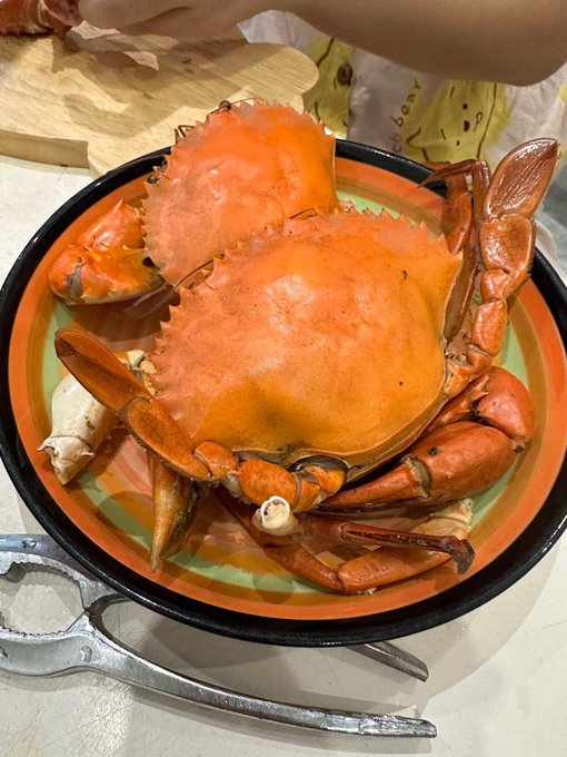 A large cooked crab served on an orange and black plate with crab-cracking tools beside it, with a person partially visible in the background.