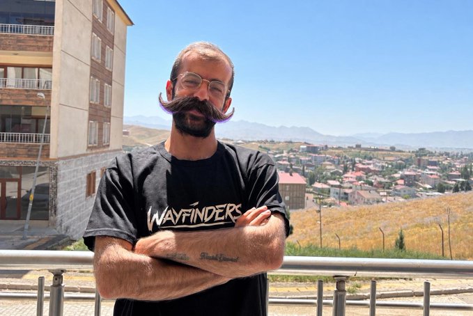 A man with a purple-tinted handlebar mustache and glasses wearing a black 'Wayfinders' t-shirt stands with crossed arms on a balcony overlooking a town with mountains in the distance.