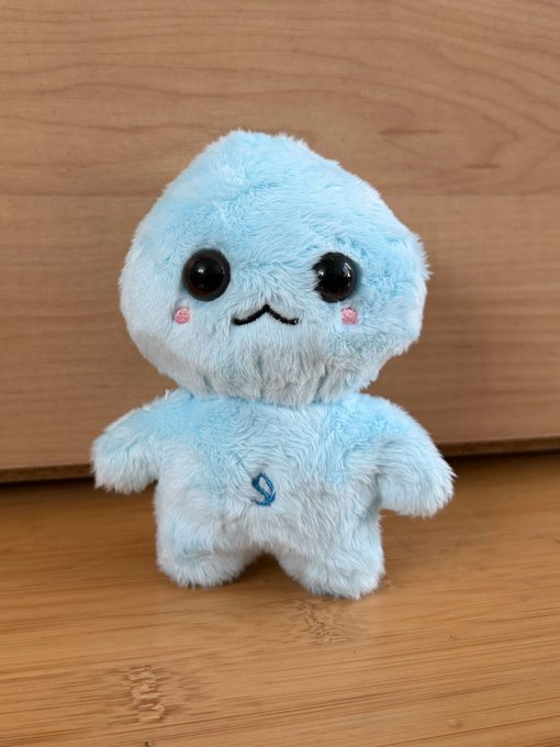 A small light blue plush toy with big black eyes, pink cheeks, and a small embroidered 'S' on its chest sits on a wooden surface.