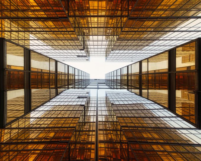 A symmetrical architectural photograph looking up through a modern glass building with golden-amber tinted windows creating a geometric pattern against the sky.