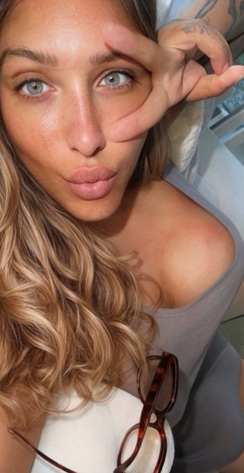 A close-up selfie of a woman with long blonde hair making an 'OK' gesture near her eye, wearing a gray top with tortoiseshell glasses tucked in.