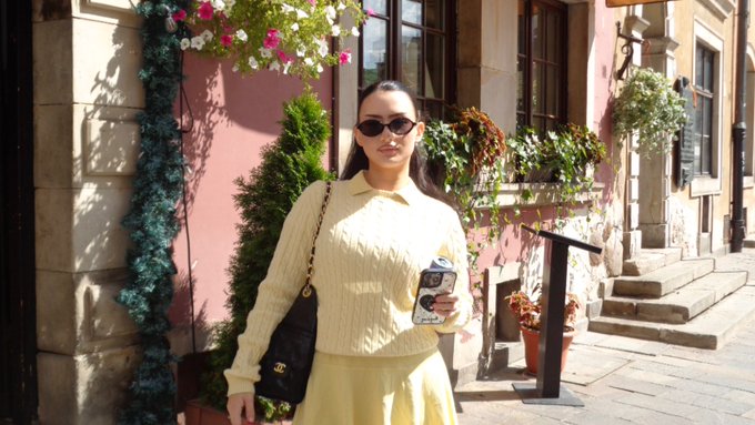 A woman wearing sunglasses, a cream sweater, and a Chanel bag stands on a sunny cobblestone street holding a phone.