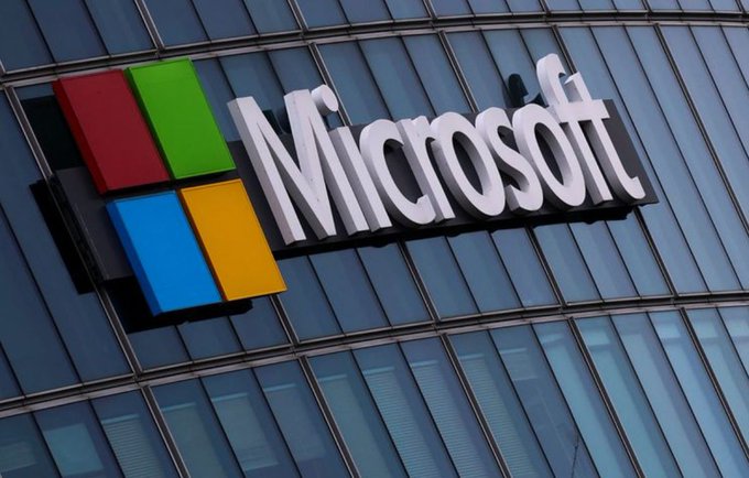 The colorful Microsoft logo is mounted on the exterior glass facade of a modern office building under bright daylight.