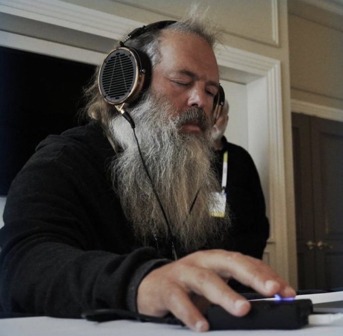 A bearded older man wearing large over-ear headphones with a distinctive grille design, appearing to be focused on listening to audio at a desk.