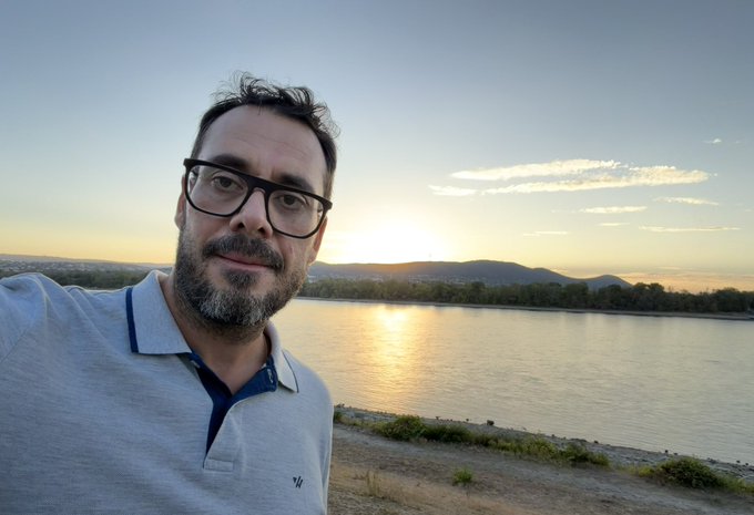 A selfie of a man with glasses and a beard wearing a light blue polo shirt, standing by a river at sunset with hills visible in the background.