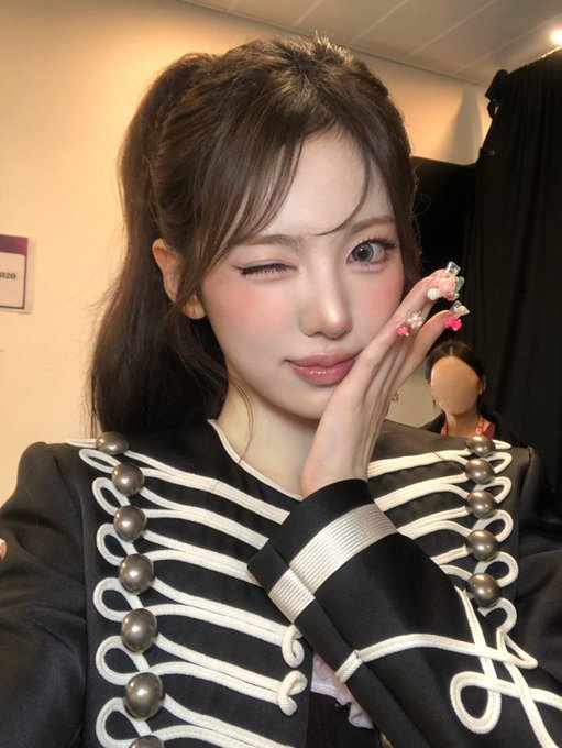 A young woman winking while resting her hand against her cheek, wearing an elaborate black outfit with white braiding and large pearl buttons, and floral-decorated nails.