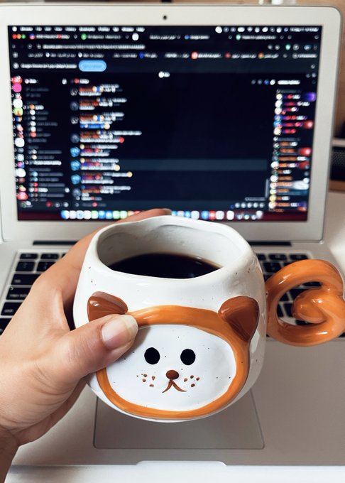 A hand holds a ceramic mug shaped like a cartoon animal face with a curled tail handle, filled with black coffee, in front of an open laptop displaying a dark-themed chat interface.