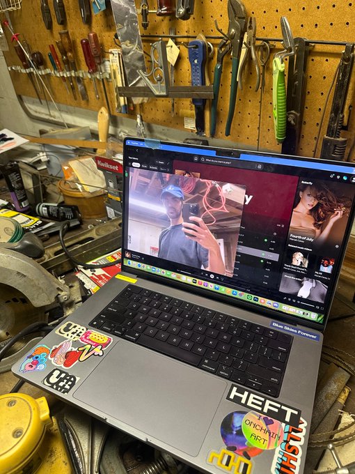 A cluttered workbench displays a MacBook Pro laptop covered in stickers, including one labeled 'HEFT,' with a pegboard of tools in the background and a video call visible on the screen.