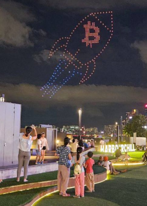 A nighttime scene of a crowd watching a drone light show forming a Bitcoin 'B' rocket in the sky, with an illuminated 'HABBO & CHILL' sign in the foreground.