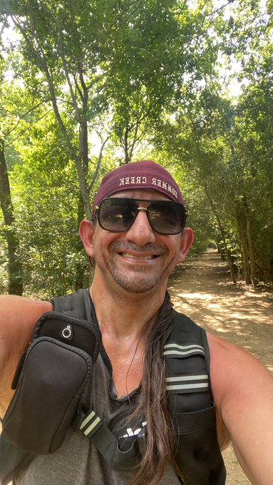A smiling man takes a selfie on a wooded hiking trail, wearing a maroon headband that reads "JIHAD JUICE" backward in the reflection, along with sunglasses and a hydration backpack, embodying the post's motivational message about self-determination and perseverance as he kicks off a Web3 weekend adventure.