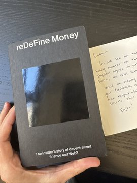 The image shows a person holding up a black book titled "Redefine Money" with a handwritten note attached to it. The note contains a message from someone else, but the specific content of the note cannot be determined from the image alone.