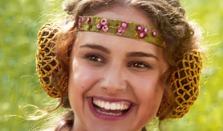 A smiling young woman with curly hair wears a colorful beaded headband and leopard-print hair snods, set against a blurred green background.
