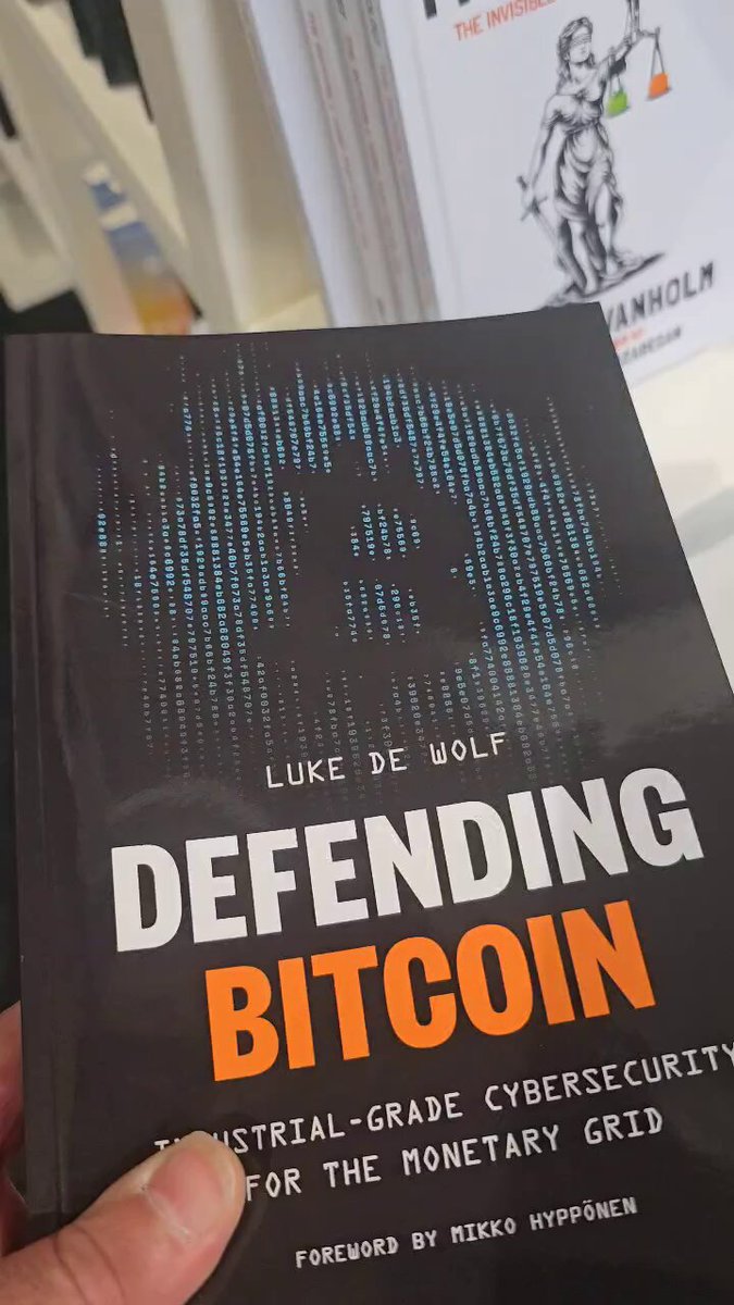 The image features a person holding up a book titled "Defending Bitcoin" with a picture of a coin on its cover, which appears to be a comic book style illustration. The book has an interesting title that suggests it might be about the history or future of Bitcoin. The person is also holding another book in their hand, possibly related to the same topic or theme as the one they are holding up.