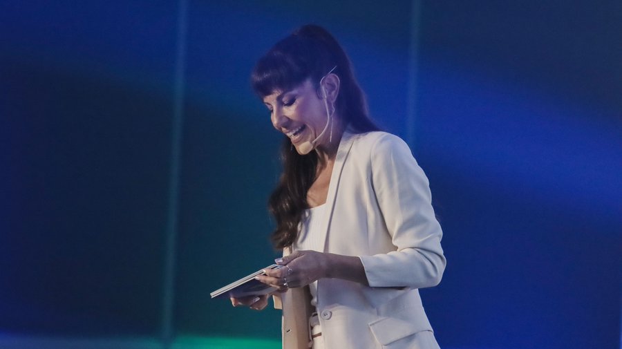 A woman in a white blazer and headset microphone smiles while reading from notes on a stage with a blue lit background.