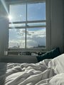 A cozy bedroom scene with white bedding, a window showing a city view with sunlight streaming in, and warm, dreamy lighting.