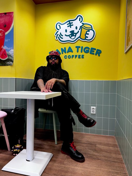 A man with long dreadlocks, sunglasses, and a red cap sits at a white table inside a 'Pana Tiger Coffee' shop, featuring a bright yellow wall with the shop's tiger logo.