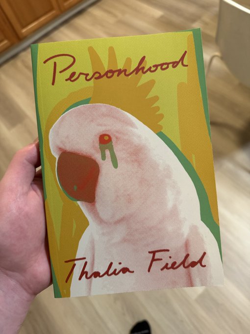 A hand holding a paperback book titled 'Personhood' by Thalia Field, featuring an illustration of a pink cockatoo on a yellow and green cover.