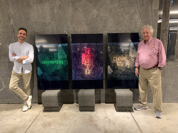 Two men pose beside three vertical displays showing "CRYPTOPUNKS," "HAROLD," and "CHARLES CSURI," referencing the Ethereum-related art collections and the computer art pioneer, with physical pixel-art sculptures placed below the screens.