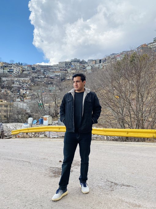 A man stands on a roadside overlooking a hillside town, wearing a dark jacket and jeans, posing for a morning greeting photo captioned "GM EVERYONE" as he asks followers to reply with "GM BACK."