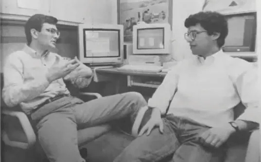A black and white archival photograph showing two men, one gesturing with his hands while the other wearing glasses listens, seated in an office with vintage computer monitors in the background.
