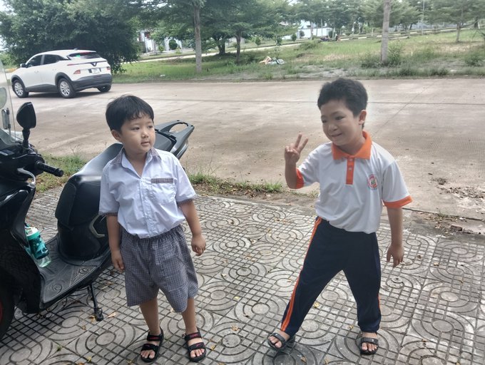 Two young boys pose outdoors on a paved surface; one wears a white button-up shirt and shorts, while the other wears an orange and white school uniform and makes a peace sign.