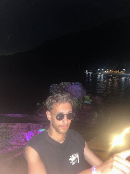 A nighttime selfie of a man wearing round glasses and a black Stüssy t-shirt, sitting by a beach or waterfront with purple-lit rocks in the background.