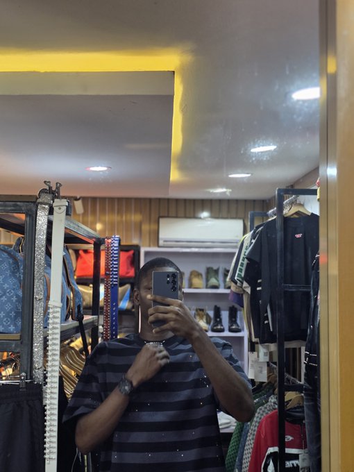 A man wearing a striped shirt takes a mirror selfie with a smartphone inside a clothing and shoe store, with shelves of garments and footwear visible in the background.