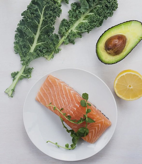 A flat-lay food photography shot featuring a raw salmon fillet garnished with watercress on a white plate, accompanied by fresh kale leaves, half an avocado with the pit, and a lemon half on a white surface.