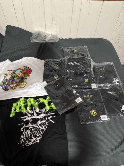 Several black and white graphic t-shirts in plastic packaging laid out on a dark couch, with one shirt displaying a green and black skull design.