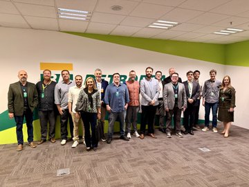 A group photo of approximately 15 people wearing lanyards standing in an office space with a green and white wall featuring the Google Cloud logo.