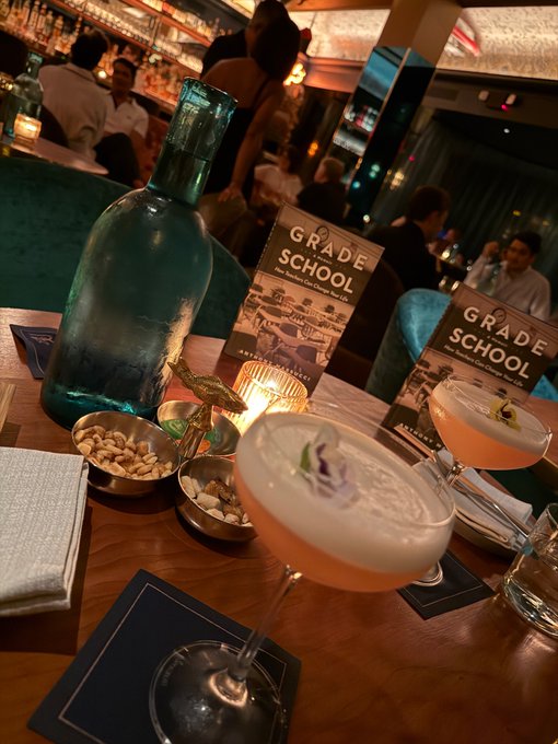 A dimly lit bar scene featuring cocktails with floral garnishes, a large teal bottle, nuts in bowls, and a card reading "GRADE SCHOOL".