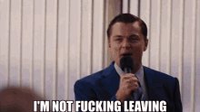 The image features a man wearing a suit, standing in front of a microphone with the caption "I'm not fucking leaving" written above him. The background shows a white wall with a window that allows light into the room. There are no other objects or people visible in the image.