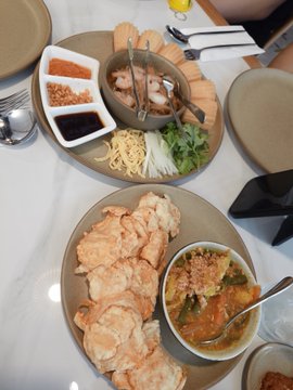 An overhead view of a restaurant meal featuring a plate of glazed meat pieces beside a bowl of curry sauce, and a divided platter with dipping sauces, garnishes, and a wooden board.