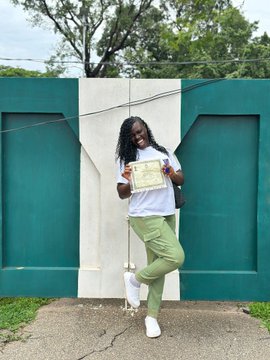 A person with long dark hair, wearing a white t-shirt, green pants, and white sneakers, poses outdoors holding a certificate in front of a green and white wall.