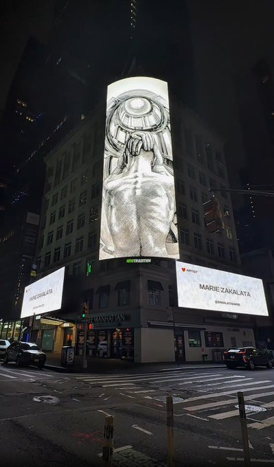 A nighttime cityscape showing illuminated billboards on a Manhattan building featuring artwork for artist 'Marie Zakalata', with the streets and traffic visible below.