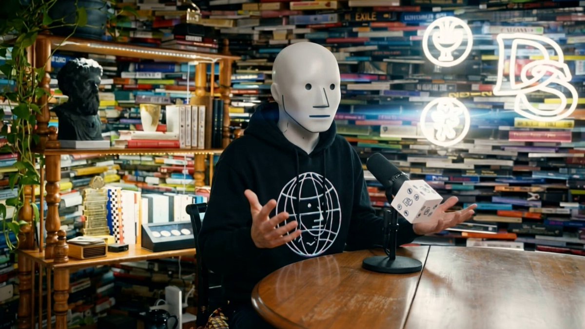 The image features a person wearing a white face mask, sitting at a table holding a microphone. They are dressed in a black hoodie and appear to be engaged in a conversation or presentation. The background of the image shows an array of bookshelves filled with various books, suggesting that they might be hosting a podcast or discussing a topic related to literature or education. The post about the podcast host's derangement suggests that this person is intentionally portraying themselves as an NPC (Non-Human) with a unique condition, possibly for artistic purposes or to create a sense of humor among their audience.