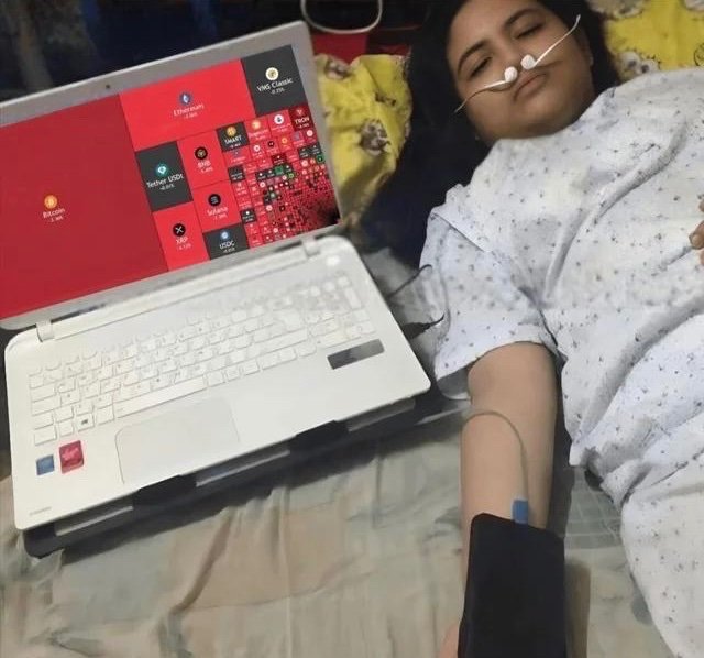A woman lies in a hospital bed wearing a nasal cannula and blood pressure cuff, with an IV line attached, while a laptop displaying cryptocurrency apps (Bitcoin, Ethereum, MATIC, VET, Polkadot, etc.) sits beside her—illustrating the post's joke that crypto holders are lying in hospitals watching their investments bleed out.