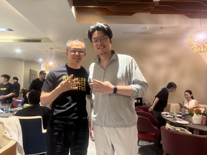 Two men posing together at an indoor restaurant or event; one wears a black 'Binance' t-shirt and the other wears a gray quarter-zip pullover.