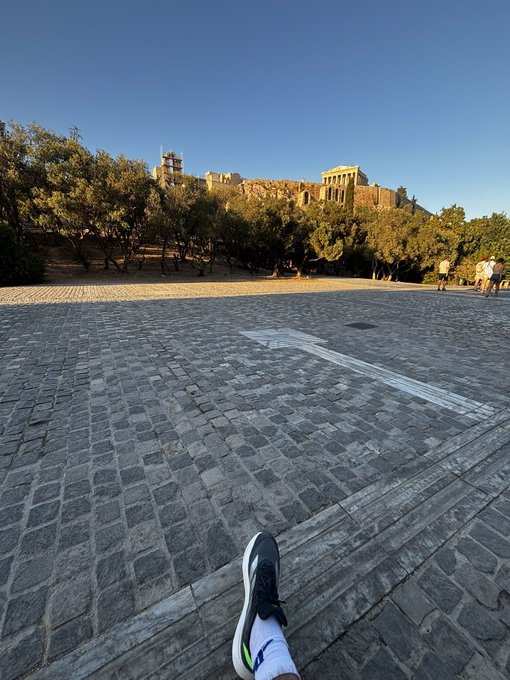 A person's foot in a black sneaker rests on a cobblestone ledge overlooking an open plaza, with an ancient stone building perched on a hill in the background.