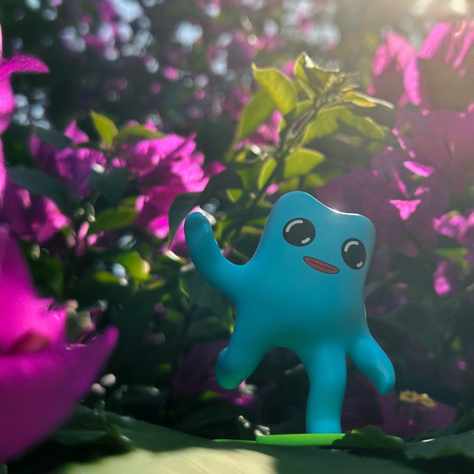 A 3D-rendered cute blue blob character standing among pink bougainvillea flowers in sunlight.
