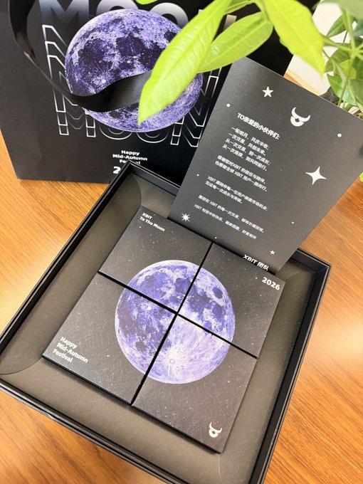 A black gift box containing four small square cards depicting the moon, accompanied by a card with Chinese text and a purple glittery moon ornament, labeled 'Happy Mid-Autumn Festival 2026'.