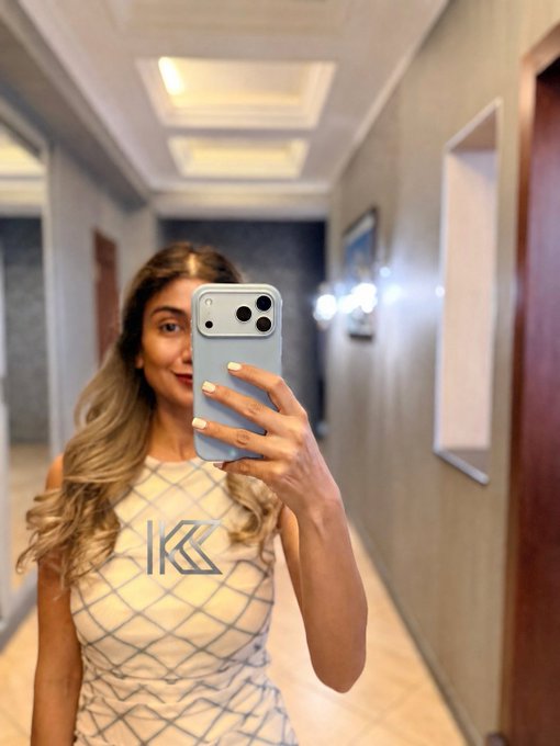 The image features a woman taking a selfie with her phone in a hallway. She is wearing a dress that has the letters "K" on it. The photo captures her reflection in a mirror as she holds up her phone, likely to take a picture of herself or share the moment with others.