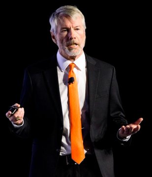 A man with gray hair and beard wearing a dark suit with a bright orange tie, gesturing with his hands while speaking, likely giving a presentation.